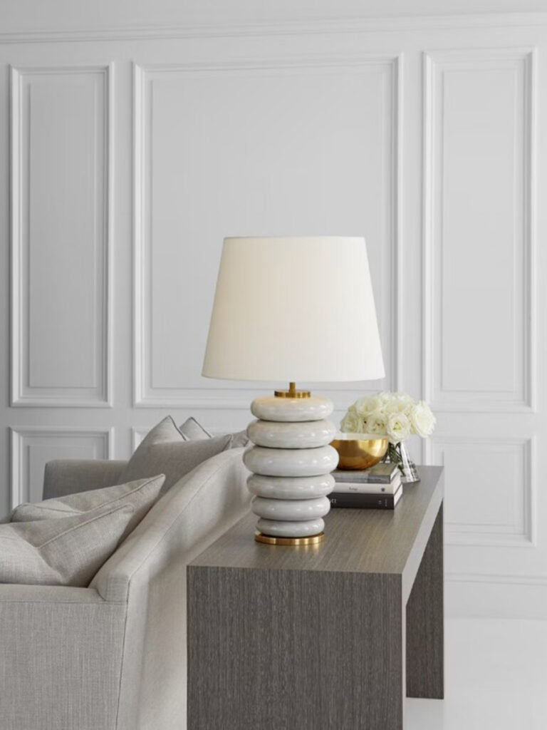 White stacked ceramic table lamp with a tapered shade on a console behind a neutral sofa.