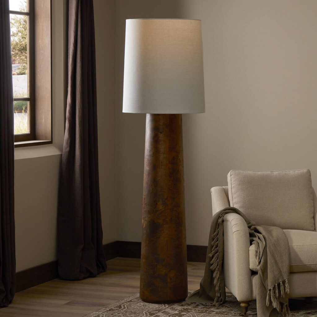 A Four Hands “Daxton” floor lamp. This tall tapered floor lamp with a warm brown base and cylindrical white shade stands beside a cream armchair.