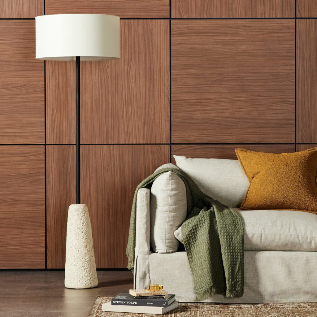 A Four Hands “Wren” floor lamp. This slim black floor lamp with a white drum shade stands beside a cream sofa and wood-paneled wall.