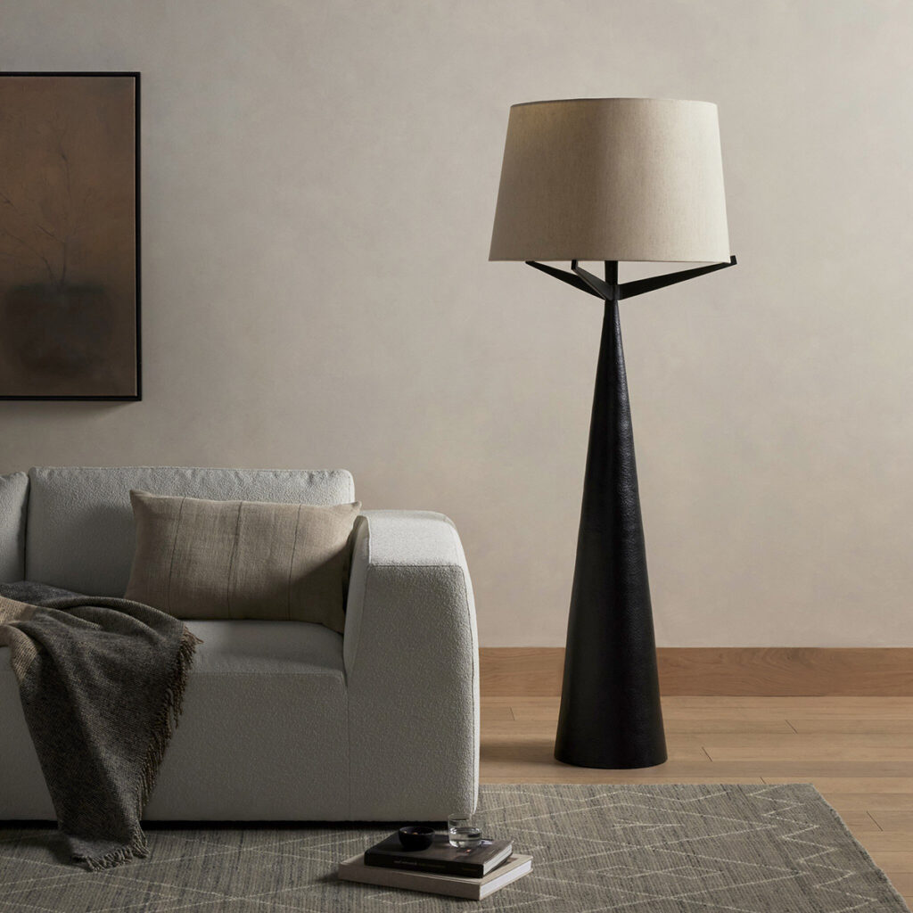 A Four Hands “Ziggy” floor lamp. This black tapered floor lamp with an angular frame and white shade stands beside a light gray sofa.
